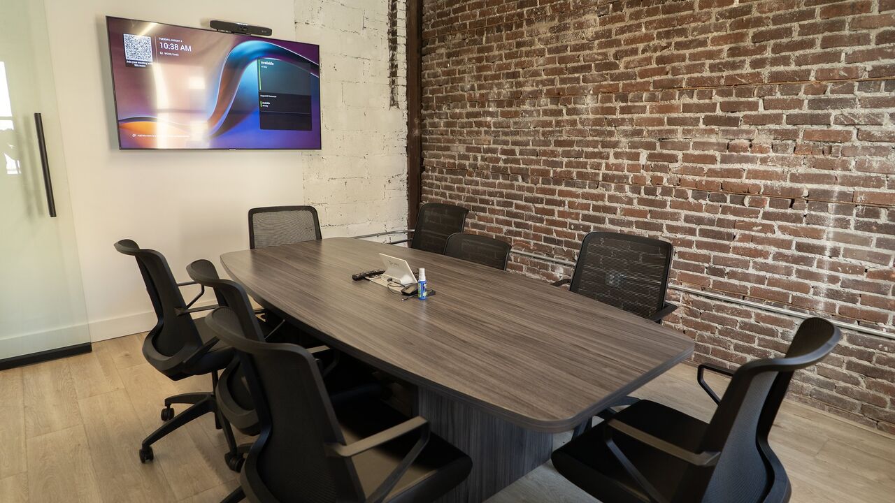 The conference room at The Node: a long meeting table ringed by mesh chairs, a wall-mounted display with a video bar, and an exposed red-brick wall