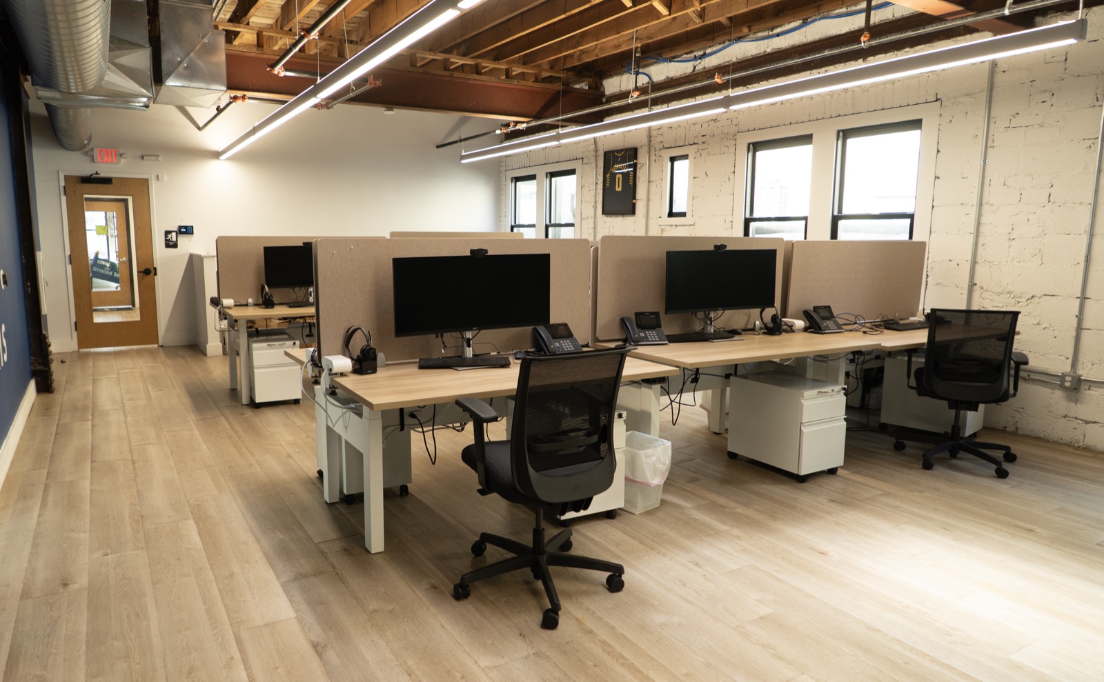 The open desk area at The Node: sit-stand desks with dual monitors along a wall of windows, under exposed timber beams