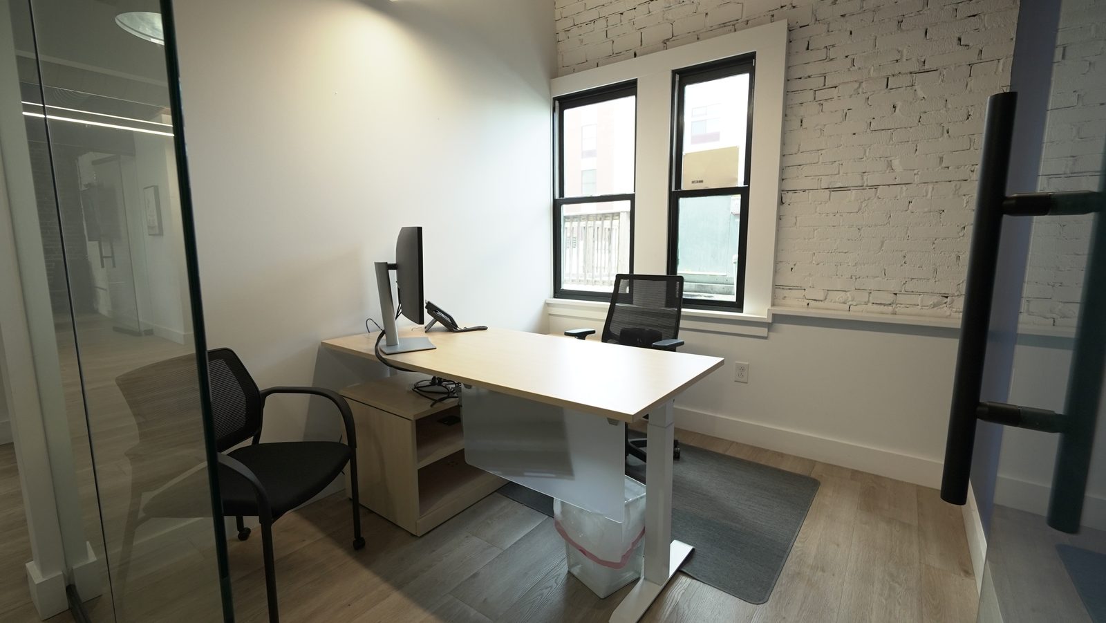 A private office at The Node seen through its open glass door: a sit-stand desk and monitor against exposed white-painted brick, with a sash window